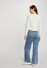 Giacca bianca corta con tessuto texturizzato, abbinata a jeans ampi blu chiaro e scarpe bianche. Vista da dietro, capelli sciolti.