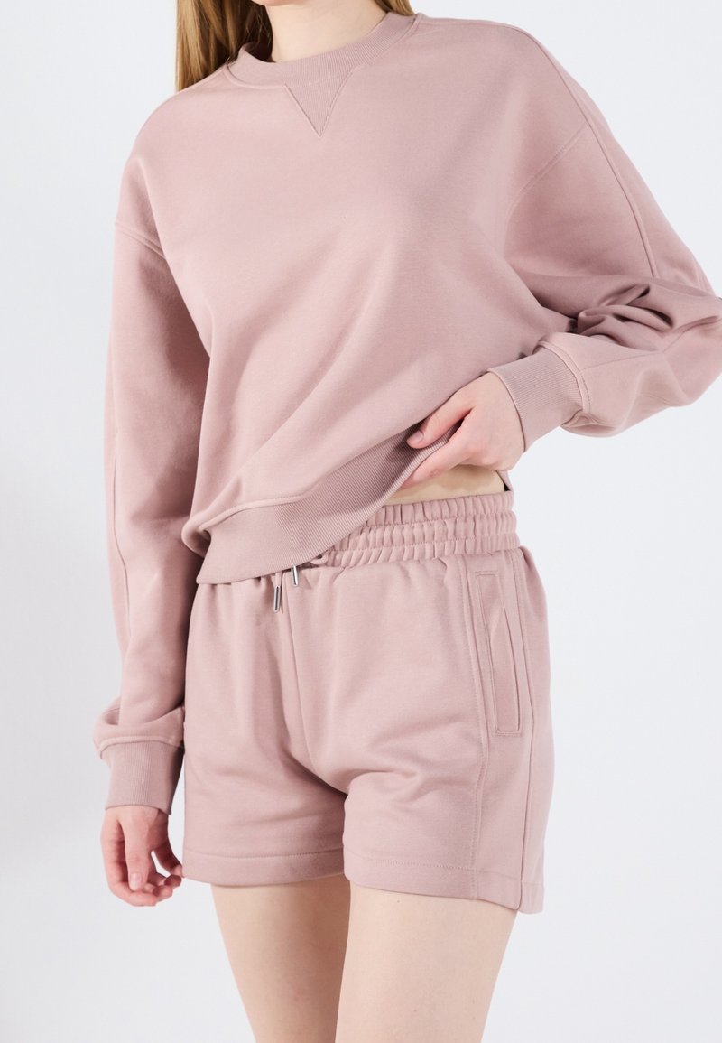 Femme portant un sweat-shirt mauve clair à manches longues et un short assorti à taille élastique, soulevant légèrement le sweat avec une main.