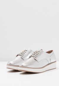 Chaussures argentées à lacets avec une tige métallique texturée, bout arrondi et semelle plateforme blanche. Design minimaliste avec des accents de couture.