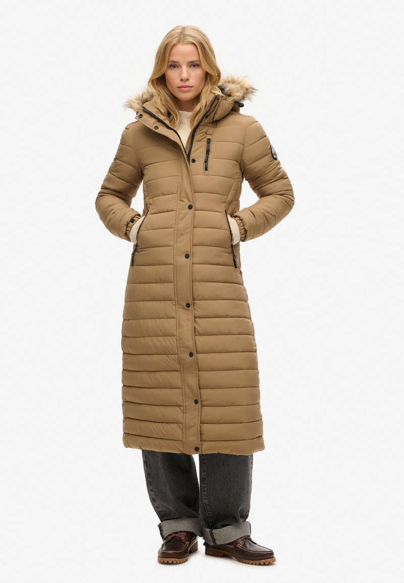 Langer, brauner Daunenmantel mit einer gefütterten Kapuze, ausgestattet mit einem gesteppten Design, Reißverschlusstaschen und Knopfverschluss. Kombiniert mit grauen Jeans und Stiefeln.