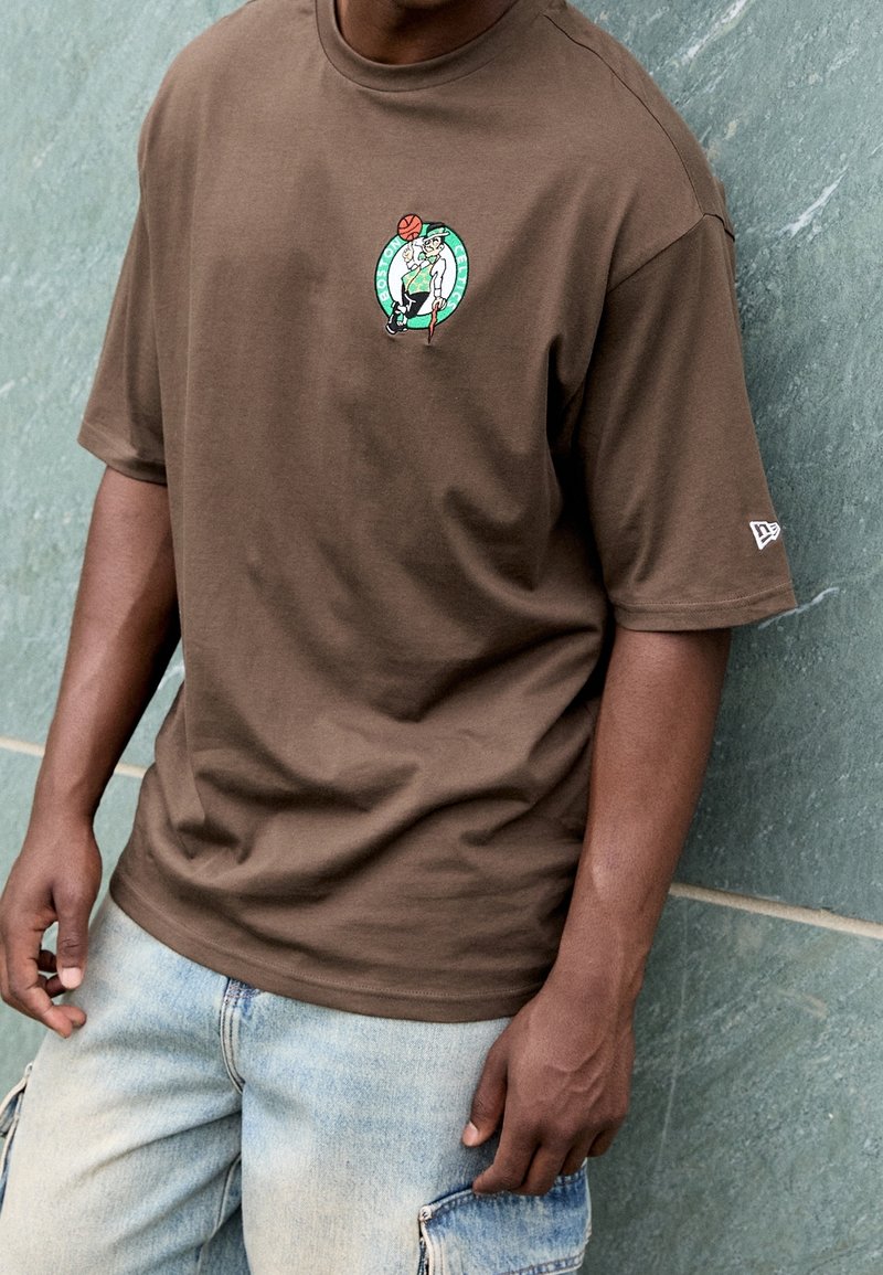 Ruskea puuvillapaita, jossa on pyöreä kaula-aukko ja lyhyet hihat. Rinnassa on brodeerattu Boston Celtics -logo, ja se on yhdistetty vaaleisiin farkkushortseihin.