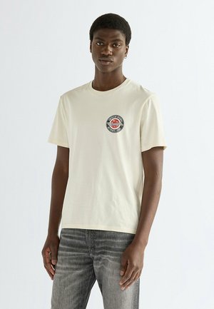 Młody mężczyzna w jasnobeżowym t-shircie z małym, okrągłym czerwono-czarnym logo na piersi i wyblakłych szarych jeansach, stojący na prostym tle.