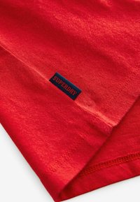 Rød bomulds T-shirt med en subtil tekstur, prydet med et blåt rektangulært label med "SUPERDRY" broderet i kanten.