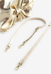 Kultainen metallinhohtoinen clutch-laukku rusettimekolla, pehmeällä pinnalla ja vetoketjusuljennalla. Mukana kaksi irrotettavaa kultahihnaa, joissa klipsikiinnikkeet.