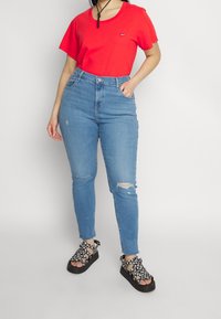 Rød kortærmet t-shirt med et lille logo, højttaljede blå jeans med slidte detaljer og mønstrede sorte slidesandaler.