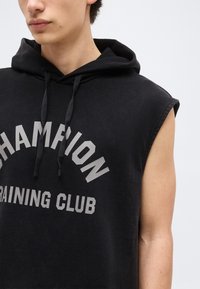 Joven con sudadera negra sin mangas con la inscripción "CHAMPION TRAINING CLUB" en gris en el frente, mirando ligeramente de lado.