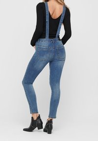 Jeanssalopette in een middelmatige wassing met verstelbare bandjes, aansluitende benen en achterzakken. Draagt bij zwarte leren enkellaarsjes met hak.