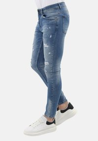 Slanke blauwe denim jeans met versleten details. Heeft een medium hoge taille en een vijf-pocket ontwerp, gecombineerd met witte sneakers.