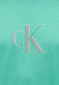 Τυρκουάζ ύφασμα με λευκό, διαγώνια ριγέ λογότυπο "cK" στο κέντρο της επιφάνειας.