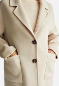 Cappotto in misto lana beige con colletto a revers, due grandi tasche frontali e due bottoni scuri. Tessuto morbido con una vestibilità su misura.
