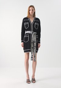 Juoda megzta suknelė su baltais akcentais, turinti dvi priekinės kišenes, V formos iškirpimą ir firminę juostelę su užrašu "KARL LAGERFELD."