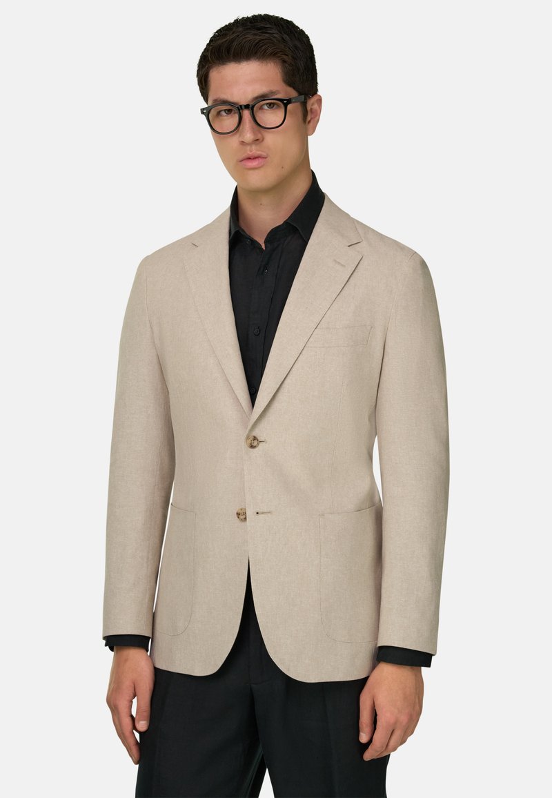 Homme portant des lunettes noires, un blazer beige à deux boutons, une chemise noire et un pantalon noir, sur un fond clair uni.