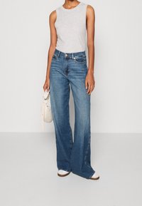 Vaqueros de mezclilla azul de pierna ancha, de cintura alta y con una textura suave; combinados con una blusa sin mangas gris y un pequeño bolso crema.