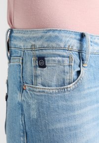 Lichtblauwe spijkerbroek met een klein achterzakje voorzien van een marineblauw label. De stof heeft een vervaagde was met subtiele textuur.