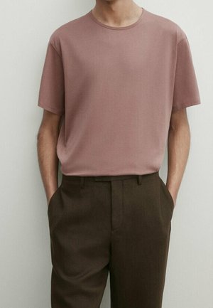 Homme portant une chemise mauve unie à manches courtes rentrée dans un pantalon taille haute marron foncé, les mains dans les poches, sur un fond neutre.