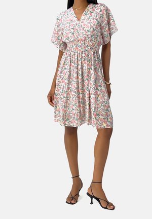 Mujer con vestido hasta la rodilla con estampado floral, mangas cortas y sandalias negras de tacón con tiras sobre fondo blanco.