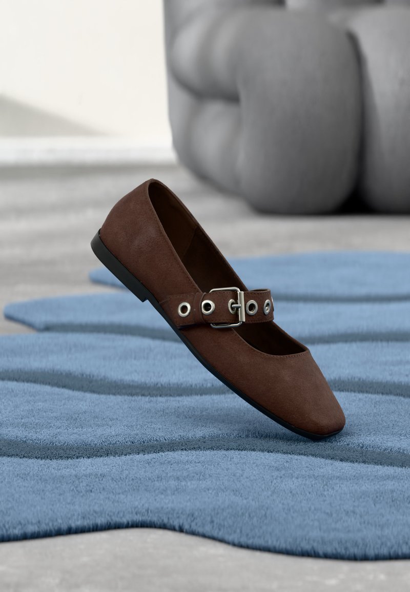 Chaussure plate en daim marron avec bride à boucle reposant sur un tapis bleu texturé, avec un meuble gris capitonné en arrière-plan.