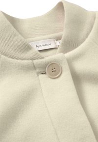 Helle beige Jacke mit glatter Textur, geripptem Kragen und runder Knopfverschluss. Etikett am Halsbereich sichtbar.