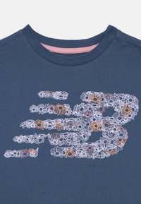 Camiseta de cuello redondo azul marino con un gran logo estilizado "NB" hecho de pequeñas flores margaritas blancas, rosas y naranjas en la parte frontal.