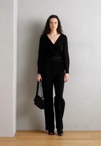 Top nero avvolgente con maniche lunghe, abbinato a pantaloni neri su misura. Gli accessori includono una piccola borsa nera. Texture lisce, design elegante.