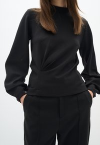 Black long-sleeve top made of smooth fabric, featuring a subtle twist detail at the waist and a high neckline. Paired with black trousers.