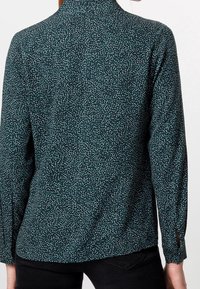 Blouse verte avec de petits pois blancs et noirs, à manches longues et col montant. Présente des fentes au niveau des poignets pour plus de liberté de mouvement.