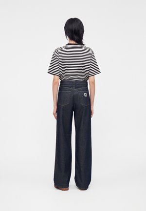 Persoon met kort donker haar, gekleed in een zwart-wit gestreept T-shirt met korte mouwen en wijde donkerblauwe jeans, van de camera afgewend.