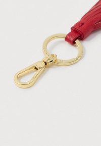 Złoty metalowy brelok z zapięciem, przymocowany do czerwonej skórzanej frędzla na białym tle.