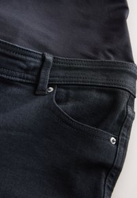 Close-up van zwarte denim jeans met voorzak, riemlus en metalen klinknagels op gestructureerde stof.