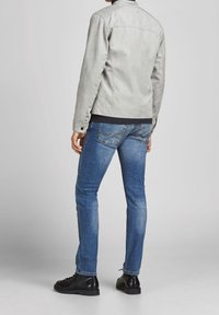 Lichtgrijze suède jas met een gestructureerd ontwerp, zwarte geribbelde kraag, gecombineerd met blauwe denim jeans en zwarte veterschoenen.