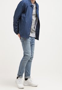 Muž v modré bundě, šedé mikině, světlých modrých džínách a bílých teniskách, stojící s rukama za zády na jednobarevném pozadí.