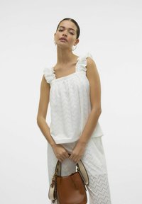 Witte blouse met ruches op de schouderbanden, voorzien van een gestructureerd zigzagpatroon. Gecombineerd met witte broeken en een leren bruine emmer tas.