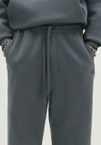 Person iført mørkegrå sweatpants og sweatshirt med hænderne i lommerne, der viser snørebåndet i livet og sølvkædearmbånd på håndleddene.