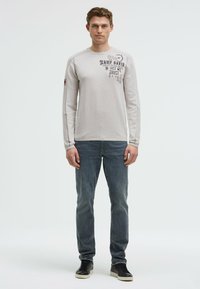 Helles graues Langarm-T-Shirt mit Grafikdruck, kombiniert mit schmalen blauen Jeans und schwarzen Sneakers, die einen lässigen, geschichteten Look zeigen.