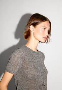 Szary dzianinowy sweter z krótkim rękawem i okrągłym dekoltem, o subtelnym połysku, noszony z długim srebrnym naszyjnikiem i zawieszką.