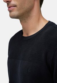 Jersey de punto oscuro que presenta una sección superior texturizada y una parte inferior suave. Detalle sutil del logo en la esquina inferior, con cuello redondo.