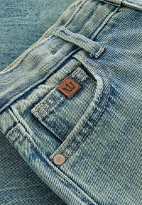 Jeans en denim de teinte bleu clair avec des coutures visibles, une petite étiquette en cuir marron et un accent en bouton métallique sur la poche avant.
