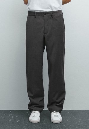 Osoba w ciemnoszarych spodniach o prostych nogawkach i białych sneakersach, stojąca na tle jednolitej jasnoszarej ściany i ciemnej podłogi.