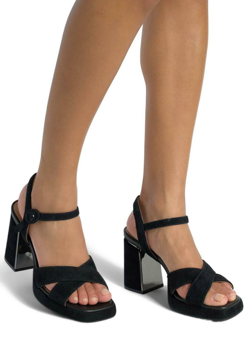 Czarne zamszowe sandały na wysokim obcasie z odkrytymi palcami, szerokimi krzyżującymi się paskami i chunky przezroczystym słupkowym obcasem. Regulowany pasek wokół kostki.