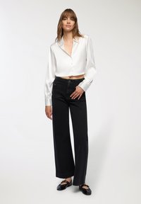 Camicia in raso bianco, tagliata, con bottoni e maniche lunghe, abbinata a jeans neri a gamba larga con vita alta. Le ballerine nere completano l'outfit.