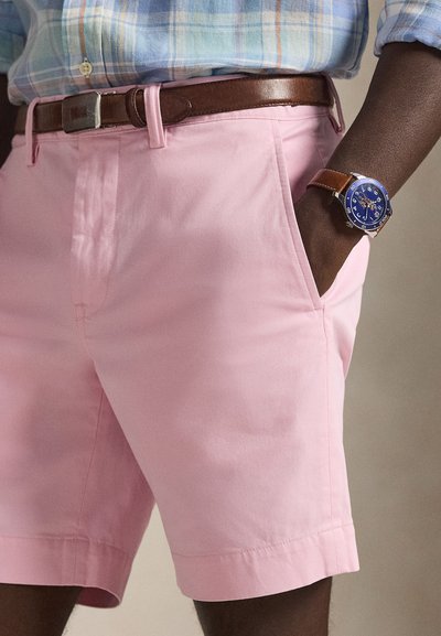 Rosa bomullsshorts med sidofickor och en slät textur. Ett brunt läderskärp och en rund blå klocka med silverdetaljer syns.