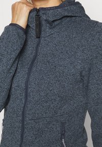 Mörkblå zip-up hoodie gjord av texturerat material med hög krage, justerbar huva och sidofickor. Har svarta dragkedjor och sömnadsdetaljer.