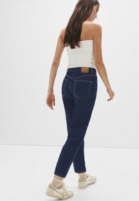 Jeans a vita alta blu scuro con un'etichetta in pelle beige, cinque tasche e cuciture a contrasto, abbinati a una maglietta bianca e sneakers chiare.