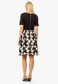 Schwarzes Kleid mit kurzen Ärmeln, tailliertem Oberteil und vollem Rock. Der Rock hat eine weiße Rosenstickerei auf dem transparenten Überzug. Mit Reißverschluss hinten.