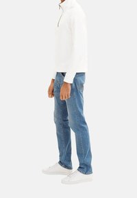 Sudadera blanca con cremallera en el cuello, jeans azules de corte slim y zapatillas blancas. La sudadera presenta una textura de canalé y los jeans tienen un diseño clásico de cinco bolsillos.