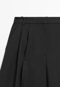 Gros plan sur un pantalon noir taille haute montrant les passants de ceinture, la poche latérale et le pli avant sur fond uni.
