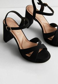 Sandali neri in suede con tacco alto, con cinturini incrociati, punta aperta e una cinghia alla caviglia regolabile. La soletta imbottita è di un colore beige chiaro.