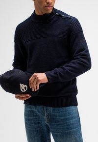 Marine gebreide trui met een hoge geribbelde kraag, voorzien van een gestructureerd bovenstuk en zijknopen. Gecombineerd met een marinekleurige pet met een logo.