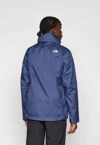 Tamsiai mėlynas nepralaidus vandeniui džemperis su aukštu apykakle, ilgomis rankovėmis ir "North Face" logotipu ant nugaros. Lygus tekstūra, prigludusi forma.