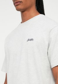 Svetlošedé bavlnené tričko s krátkymi rukávmi a okrúhlym výstrihom, na hrudníku s tmavomodrým vyšitým logom "JACKER".